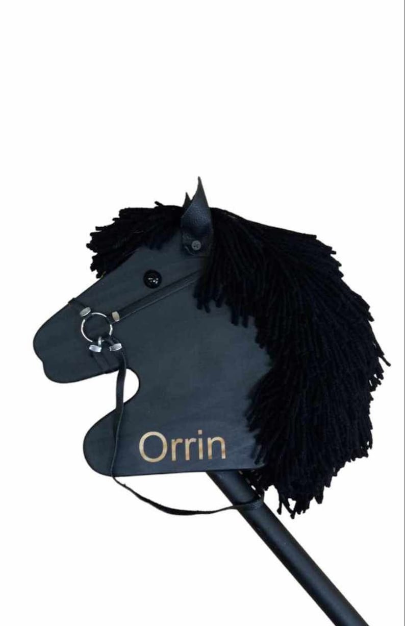 May include: Black wooden stick horse toy with black yarn mane and tail. The horse has a black leather bridle with a silver bit and a gold name tag that says "Orrin".