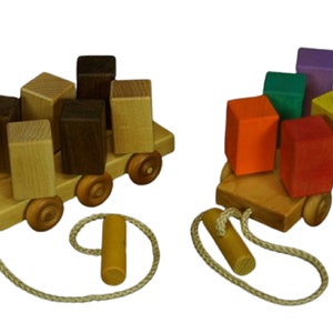 May include: Two wooden toy trucks with colorful blocks stacked on top. The trucks have wheels and a rope attached to pull them. One truck has brown and natural wood blocks, the other has orange, red, green, yellow, purple, and blue blocks.
