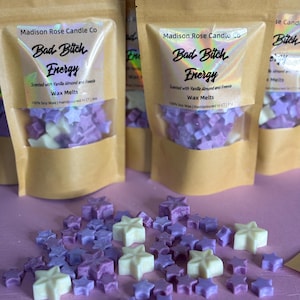 May include: Several brown paper pouches filled with star-shaped wax melts in shades of purple and white. The labels read Madison Rose Candle Co, Bad Bitch Energy, Scented with Vanilla Almond and Freesia, Wax Melts, 100% Soy Wax, Handpoured in CT.