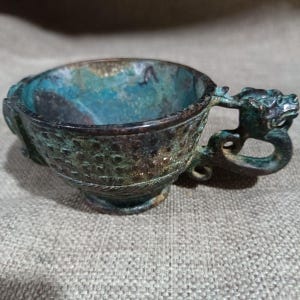 Puede incluir: Taza antigua de bronce con asa en forma de dragón. La taza tiene un exterior texturizado y un interior turquesa. El asa está intrincadamente diseñada con una cabeza de dragón. Probablemente un artefacto antiguo.