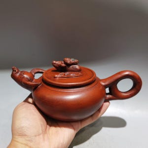 Könnte beinhalten: Eine rotbraune Tonteekanne mit einem skulptierten Tierkopf am Ausguss und einem kreisförmigen Griff. Der Deckel ist mit einer detaillierten Tierfigur verziert, die ein glattes, mattes Finish und kunstvolle Handwerkskunst aufweist.