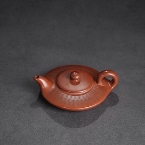 May include: A low-profile, reddish-brown clay teapot with a flat, circular body and a matching lid featuring a small knob. The surface of the teapot is decorated with a band of intricate, dark-coloured Chinese characters engraved around the centre.