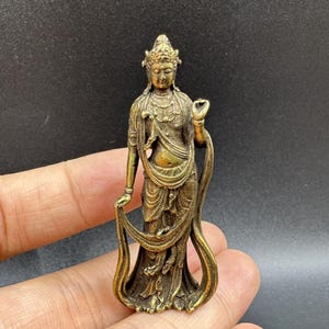 Può includere: Una piccola statuetta in ottone in stile antico raffigurante una divinità in piedi con una corona ornata e vesti fluenti. La figura ha una mano sollevata in un gesto e l'altra drappeggiata nel tessuto, con una finitura metallica dorata.