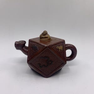 May include: A dark brown, hexagonal teapot featuring a textured surface with embossed animal figures. The spout is shaped like a feline head, the handle is curved, and the lid is topped with a small, light-colored monkey figurine.