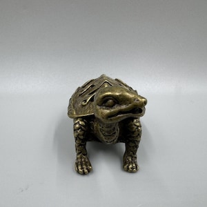 Könnte beinhalten: Eine kleine Schildkrötenfigur aus Messing im Antik-Stil mit einer texturierten, verwitterten Oberfläche. Die Schildkröte hat einen detaillierten, gemusterten Panzer, einen geformten Kopf mit definierten Augen und Mund sowie vier Beine mit Krallen.