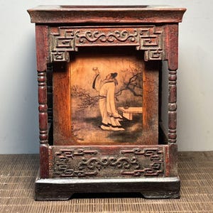 Puede incluir: Una caja rectangular de madera con acabado marrón oscuro, que presenta patrones geométricos tallados en los bordes superior e inferior. El panel central muestra una ilustración pintada de una figura con túnicas tradicionales sosteniendo una copa.