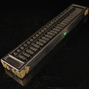 Op de afbeelding: Een rechthoekige abacus van donker hout met meerdere rijen verschuifbare kralen, versierd met sierlijke goudkleurige metalen hoekplaten en een enkele ovale groene steen ingelegd in het zijframe, een traditioneel rekeninstrument.