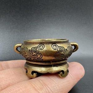 May include: A small, round, metallic bronze-toned incense burner featuring two side handles and intricate, embossed swirling patterns along the exterior. The vessel rests on a decorative, three-legged base with curved, ornate cut-out details.