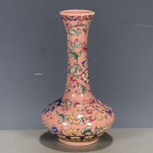May include: A tall, slender ceramic vase with a light pink base colour, featuring a repeating pattern of colourful floral motifs and swirling green vines. The design includes small blue, yellow, and red blossoms distributed across the surface.