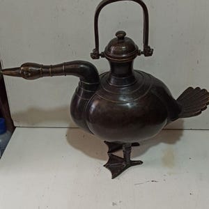 May include: A bronze duck-shaped vessel with a handle, beak, and tail feathers. The vessel has a round body and stands on webbed feet. The dark brown color gives it an antique appearance. The handle is arched and attached to the top of the vessel.