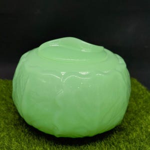 May include: A round, light green jadeite glass bowl with a matching lid. The exterior surface features a molded, textured pattern resembling overlapping cabbage leaves, with a smooth, curved handle integrated into the center of the lid.