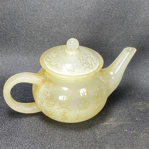 Peut inclure: Une théière translucide blanc cassé avec un corps arrondi, une anse courbée et un bec verseur. La surface de la théière et de son couvercle est ornée de caractères chinois gravés et de motifs floraux, présentant une finition lisse et polie.
