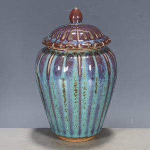 Pode incluir: Um pote de cerâmica com tampa apresentando uma textura canelada vertical. O esmalte exibe uma mistura de tons turquesa, azul-petróleo e roxo profundo com acabamento brilhante. A tampa possui um pequeno puxador arredondado no topo.