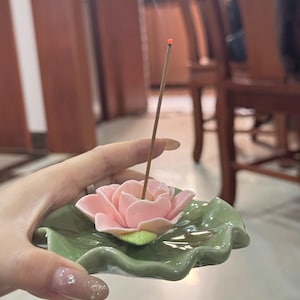 May include: A pink lotus flower incense holder with a green leaf-shaped base. The incense holder is made of ceramic and has a glossy finish. An incense stick is lit and placed in the center of the flower.