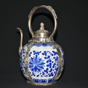 May include: A round ceramic teapot featuring a white base with blue floral patterns. The teapot is encased in silver-toned metal framework with ornate lion head accents, a curved spout, a high arched handle, and a lid topped with a small figure.