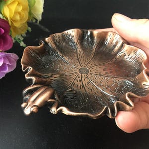Pode incluir: Um prato decorativo de cor bronze em forma de folha de lótus, com uma figura detalhada de rã na lateral. O prato tem uma superfície texturizada e um design circular central.