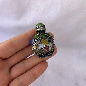 Puede incluir: Una pequeña botella de rapé de metal ornamentada con un acabado oscuro y texturizado con intrincados patrones de filigrana plateada. La botella está decorada con piedras cabujón verdes y azules incrustadas y un motivo central de insecto esmaltado.