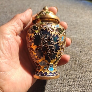 Può includere: Un piccolo vaso in metallo dorato con coperchio abbinato, caratterizzato da un intricato lavoro di smalto in stile cloisonné nei toni del blu scuro, azzurro, verde e bianco, con un motivo floreale e contorni in filo d'oro in rilievo.