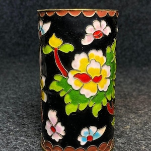 Può includere: Un vaso cilindrico in metallo con uno sfondo in smalto nero decorato con colorati motivi floreali cloisonné. Il design include grandi fiori gialli e bianchi con centri rossi, foglie verdi e fiori più piccoli di colore rosa e blu.