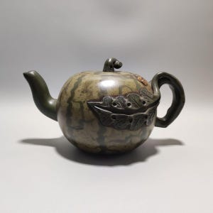 May include: A round ceramic teapot with a muted green and brown mottled finish resembling a watermelon. It features a curved spout, a handle, a small lid knob, and a decorative relief of a textured leaf on the side with small circular cutouts.