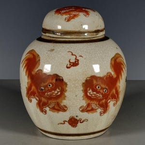 May include: A ceramic ginger jar with a cream-colored crackle glaze finish. The jar features orange painted lion-like creatures on the body and lid, accented by small decorative motifs. It has a rounded shape with a matching removable lid.