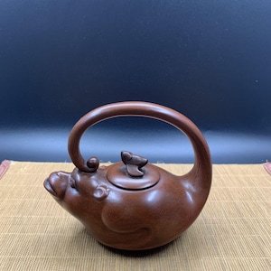 May include: A brown clay teapot shaped like a stylized animal, featuring a curved handle that arches over the top, a small lid with a sculpted finial, and a spout integrated into the animal's head design with visible facial features.