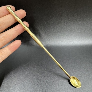 Puede incluir: Una cuchara de bar dorada con un mango largo y delgado y una pequeña cuchara ovalada en el extremo. El mango tiene un diseño decorativo con una parte superior redondeada. La cuchara está diseñada para mezclar cócteles y otras bebidas.