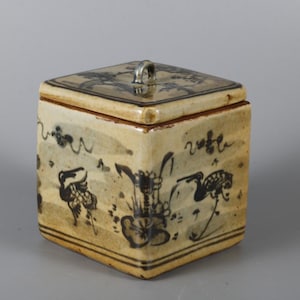 May include: A square ceramic container with a lid featuring a small handle. The piece has a cream-colored glaze with dark blue hand-painted illustrations of birds and floral motifs, finished with horizontal line borders at the top and bottom edges.