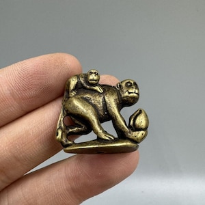 Peut inclure: Petite figurine de singe de couleur bronze. La sculpture représente un grand singe portant un petit singe sur son dos, tenant un fruit. La figurine a une surface détaillée et texturée et est probablement en métal.