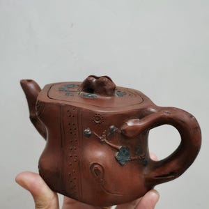 May include: A brown clay teapot featuring a square body with a textured surface, a curved handle, and a spout. The lid is adorned with small blue floral accents and a central knob, while the body displays vertical dotted lines and blue leaf motifs.