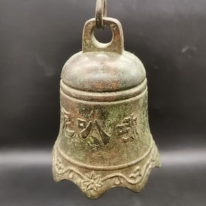 May include: A weathered bronze-toned metal bell featuring a loop at the top for hanging. The surface displays a dark, oxidized patina with embossed Japanese characters and a decorative scalloped bottom edge adorned with small floral motifs.