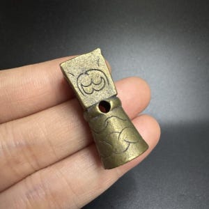 Puede incluir: Pequeño colgante antiguo de color bronce. La parte superior rectangular tiene un diseño tallado y un agujero circular. La parte inferior cilíndrica tiene un patrón texturizado. El colgante se sostiene en una mano.