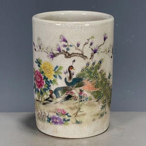 May include: A cylindrical ceramic brush pot with a cream-coloured crackle glaze finish. The exterior features a detailed painted scene of two peacocks surrounded by vibrant yellow, pink, and purple flowers and green foliage under a tree branch.