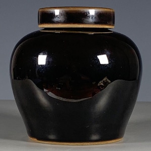 Peut inclure: Un pot en céramique brun foncé brillant avec un corps arrondi et un couvercle assorti. La glaçure présente une finition lisse et réfléchissante avec de subtils reflets brun clair le long du bord du couvercle et de la base du pot.
