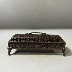 Puede incluir: Una caja decorativa rectangular de color bronce con tallas ornamentadas y un asa. La caja tiene una parte superior calada y está sostenida por cuatro patas curvas. Los lados presentan patrones en relieve detallados.