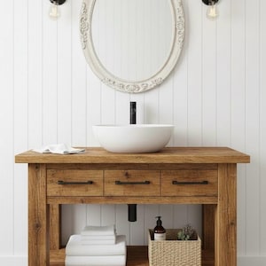 May include: A rustic wooden bathroom vanity with a white oval sink and three drawers. A decorative white framed mirror hangs above the sink. The vanity has a lower shelf with folded white towels and a woven basket. Two black sconces with Edison bulbs are mounted on the wall.
