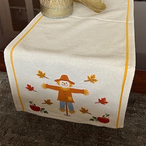 May include: A cream-coloured fabric table runner featuring a yellow border and an embroidered scarecrow surrounded by autumn leaves and pumpkins. The runner is decorated with a gold-patterned glass vase, a gold leaf-shaped dish, and glass ornaments.