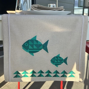 Puede incluir: Un camino de mesa rectangular de color beige con dos peces bordados en tonos verde azulado y verde oscuro, compuestos por patrones triangulares geométricos, con una fila de triángulos verdes en el borde inferior y bordes ribeteados en blanco.