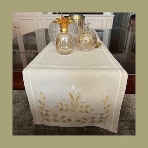 May include: A beige table runner with embroidered floral and leaf designs. The runner is decorated with white tulips and gold-colored leaves. Decorative glass pineapple and orb-shaped objects sit on the runner.