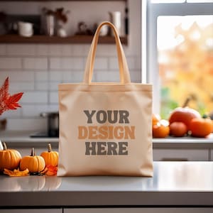 May include: A beige tote bag with the text "YOUR DESIGN HERE" in brown and gray. The bag has a rectangular shape with a handle. The background includes pumpkins and fall foliage, suggesting a seasonal theme.