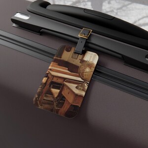 Può includere: Un'etichetta per bagagli con una scena di classe vintage con una scrivania in legno, una sedia e un mappamondo. L'etichetta è attaccata a una maniglia di valigia nera con una fibbia color oro. L'etichetta ha un cinturino nero.