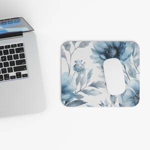 May include: A white mousepad with a watercolor floral design in shades of blue and gray. A white computer mouse rests on the mousepad. A laptop keyboard is visible in the upper left corner.