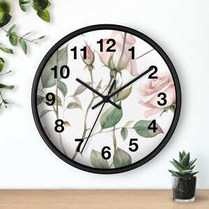 May include: A round wall clock with a black frame and a white face featuring a watercolour illustration of pale pink roses, green leaves, and stems. The clock has black hour, minute, and second hands with large, bold black numbers from 1 to 12.