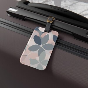 May include: A rectangular luggage tag featuring a repeating floral-inspired pattern in shades of dusty blue, slate gray, and soft blush pink. The tag is attached to a dark brown suitcase handle by a black strap with a gold-toned metal buckle.