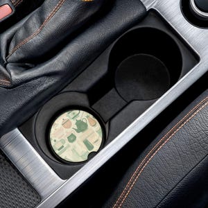 Pode incluir: Uma base de copo redonda para carro com um padrão de chá de bules, xícaras e outros acessórios de chá em tons de verde, pêssego e creme. A base de copo está em um porta-copos de carro.