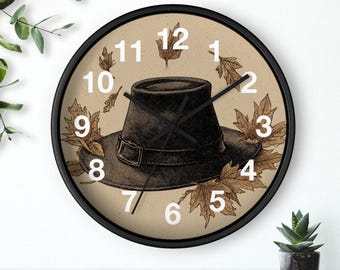 May include: A round wall clock with a black frame. The clock face features a vintage illustration of a black pilgrim hat, surrounded by brown autumn leaves. White numerals mark the hours.