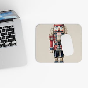 May include: A rectangular mousepad featuring a detailed illustration of a traditional nutcracker soldier. The nutcracker is dressed in a red and blue uniform with gold accents. A white computer mouse rests on the right side of the mousepad.