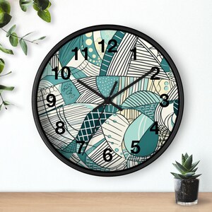 Könnte beinhalten: Eine runde Wanduhr mit schwarzem Rahmen und weißem Zifferblatt, das ein abstraktes Muster aus Linien, Punkten und geometrischen Formen in Petrol, Dunkelgrün und Creme zeigt. Die Uhr hat schwarze Zeiger und große schwarze Ziffern.
