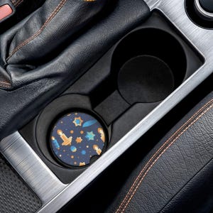 Pode incluir: Um porta-copos de carro azul marinho com um padrão de velas, estrelas e outras formas em laranja, azul e amarelo. O porta-copos está colocado no porta-copos de um carro, rodeado por couro preto e acabamentos prateados.