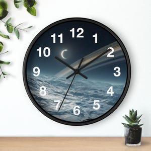 May include: A round wall clock with a black frame and a face featuring a space-themed image of a planet surface with rings and a crescent moon in a dark sky. The clock has white numbers from 1 to 12 and black hour, minute, and second hands.